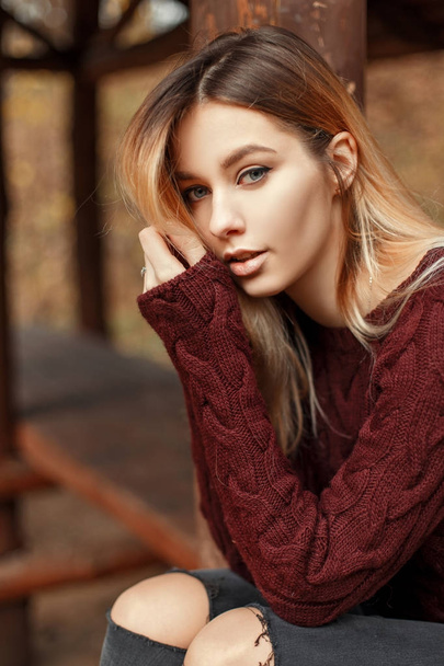 Fashion portrait of a beautiful young blonde girl in a stylish sweater outdoors on the autumn day sits near wooden lodge - Foto, Imagem