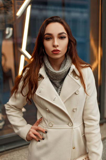 Стильная молодая девушка в осеннем трикотажном пальто позирует на улице перед магазином
 - Фото, изображение