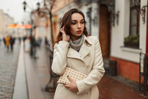 Fashionable beautiful young woman in a stylish coat with a fashionable bag walking outdoors in the city - Foto, imagen