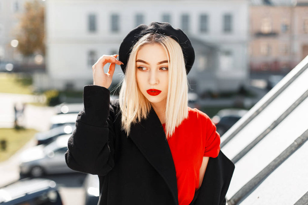 Beautiful fashionable young woman with red lips in a black coat and black beret in the street on a sunny day - Photo, Image