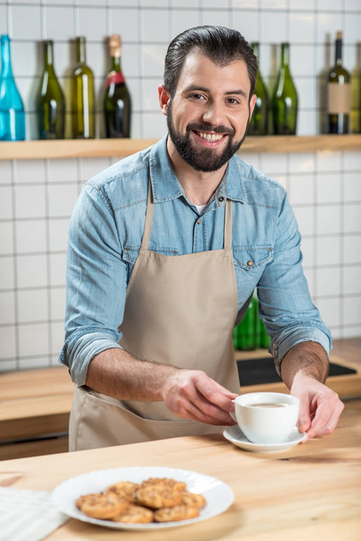 Έμπειρος σερβιτόρος βάζοντας ένα φλιτζάνι του καφέ σε ένα τραπέζι, ενώ χαμογελώντας - Φωτογραφία, εικόνα