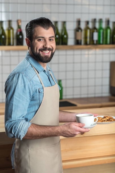 Handsome friendly waiter smiling while holding a cup of coffee and cookies - Foto, immagini