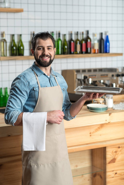 Handsome bearded waiter holding a tray and smiling cheerfully - 写真・画像