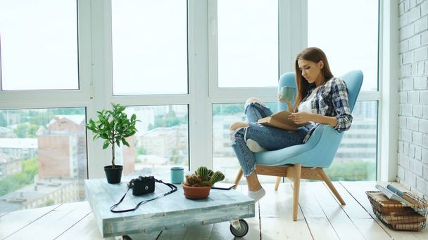 Young attractive woman read book and drink coffee sitting on balcony in modern loft apartment - Valokuva, kuva