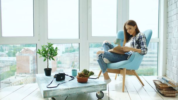 Young attractive woman read book and drink coffee sitting on balcony in modern loft apartment - Photo, image