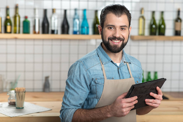 Cheerful bearded waiter holding a modern tablet and smiling - Foto, Imagem
