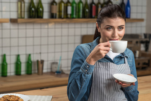 Cute smiling waitress standing at the bar counter and drinking tasty tea - Fotó, kép