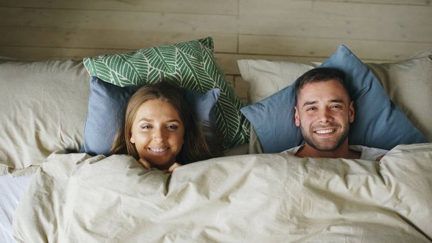 Top view of smiling couple having fun in bed hiding under blanket and looking into camera - Foto, Bild