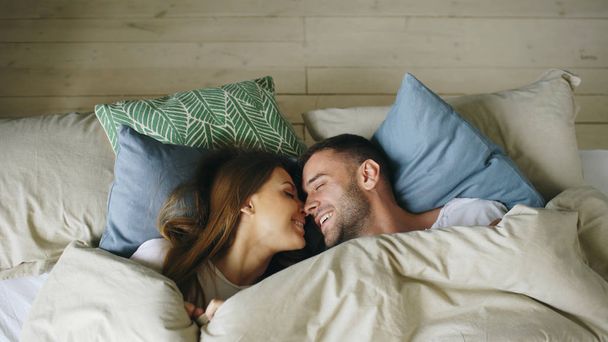 Top view of couple having fun in bed lying under blanket looking into camera and kissing at home - Foto, afbeelding