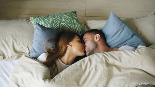 Top view of couple having fun in bed lying under blanket looking into camera and kissing at home - Foto, Imagem
