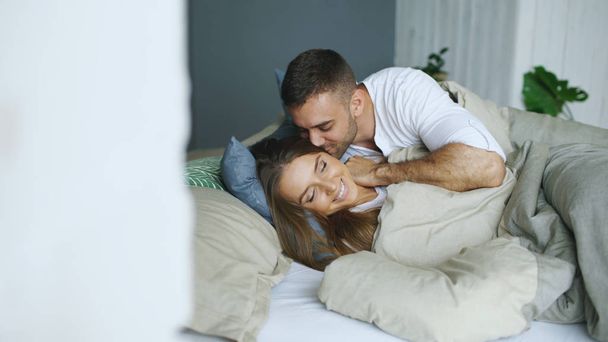 Young beautiful and loving couple wake up at the morning. Attractive man kiss and hug his wife in bed - Photo, image