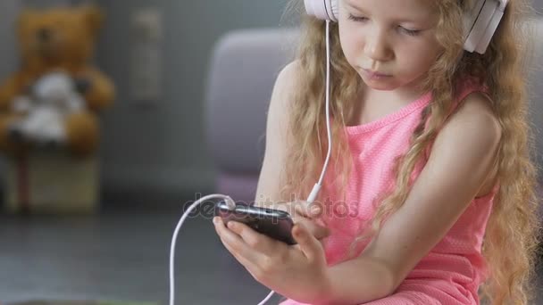 Funny female child listening to music in headset, holding smartphone, closeup - 映像、動画