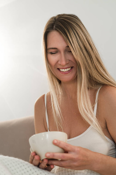 schöne Frau mit langen blonden Haaren wacht auf und sitzt mit einer Tasse Tee oder Kaffee im Bett. Morgen träumen und zu Hause entspannen - Foto, Bild