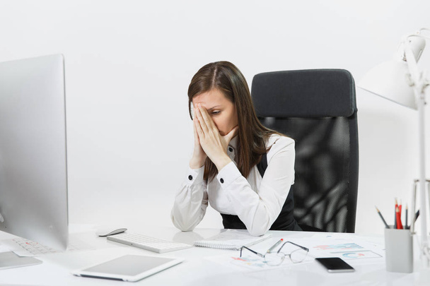Уставшая от недоумения и стресса каштановые волосы деловая женщина в костюме сидит за столом, закрывая лицо руками, работает за современным компьютером с документами в светлом офисе на белом фоне
 - Фото, изображение