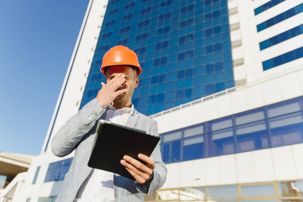 Moe zakenman in pak en beschermende constructie oranje helm met tablet covers gezicht met zijn hand tegen het gebouw in blauwe kleur. Ingenieur in de helm voor de veiligheid van de werknemers op kantoorgebouwen. - Foto, afbeelding