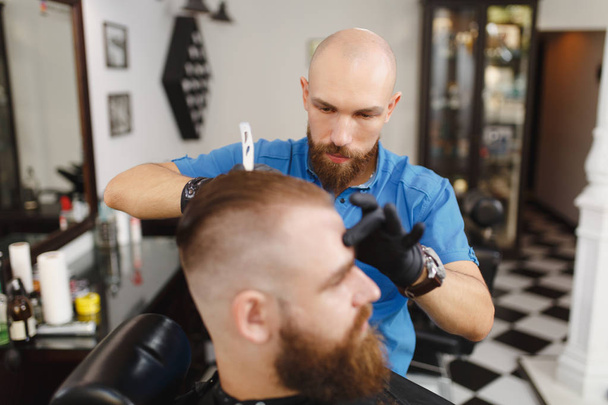 Мужчина профессиональный парикмахер, обслуживающий клиента, бреющий толстую большую бороду бритвой. Рыжий красивый стильный молодой человек с короткой стрижкой, черная накидка. Светло-белая парикмахерская
 - Фото, изображение