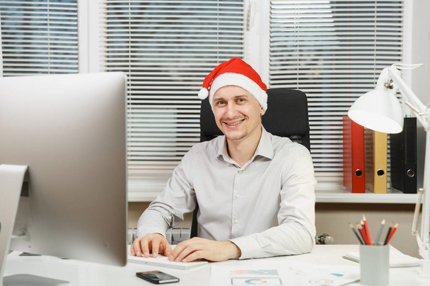 Handsome smiling business man in shirt and red Christmas hat sitting at the desk, working at computer with modern monitor, with documents in light office, hands on the keyboard, on window background. - Fotografie, Obrázek