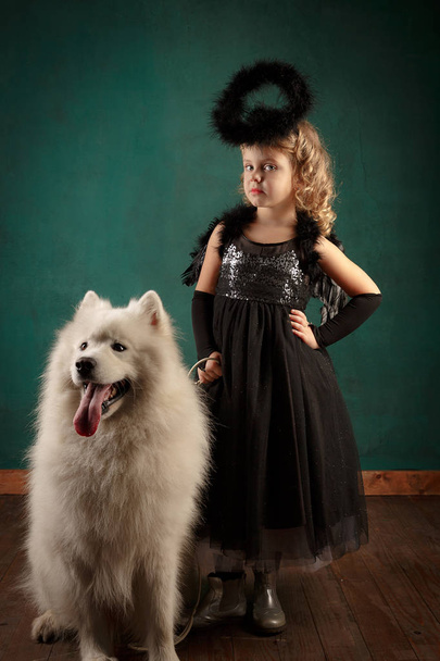 Composição de Ano Novo no estúdio. Loira bonito pouco em um traje de anjo preto com um grande cão branco. Ano do cão, Humor
 - Foto, Imagem