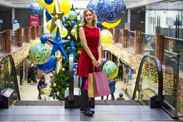 Young beautiful girl enjoys shopping in a shopping center with bright packages on the background of a Christmas tree - Photo, Image