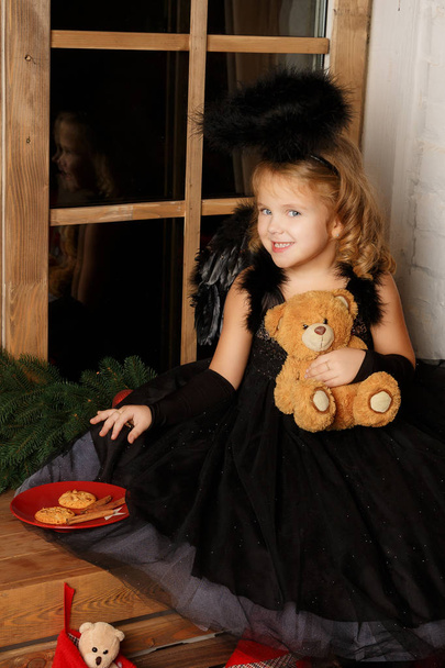 クリスマス シーン。窓の近くのサンタのクッキーを盗んで黒い天使衣装で小さなブロンドの女の子の肖像画. - 写真・画像