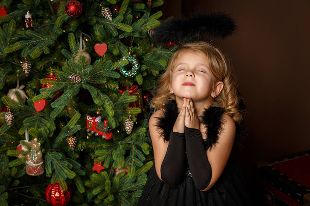 Reza a una niña con un disfraz de ángel negro, buscando con esperanza la paz. Feliz infancia y paz. Navidad, Año Nuevo
 - Foto, Imagen