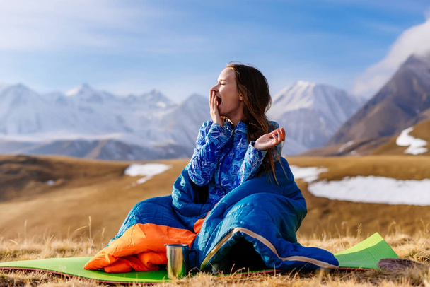 a young girl loves to travel, sits in a sleeping bag on the ground, yawns, against the background of the Caucasian ridge - Photo, image