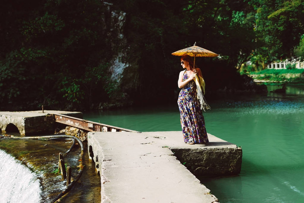 red-haired woman in a long dress walking through the park in Asia, holding a Chinese umbrella - Fotó, kép