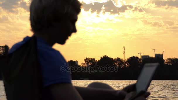 Ο άνθρωπος κάθεται σε ένα διπλώνοντας καρέκλα και κοιτάζει του Tablet pc στο ηλιοβασίλεμα εκτός εστίασης - Πλάνα, βίντεο