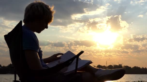明るい男座っている読み取り、日没で川岸に彼のノートを書き込みます - 映像、動画