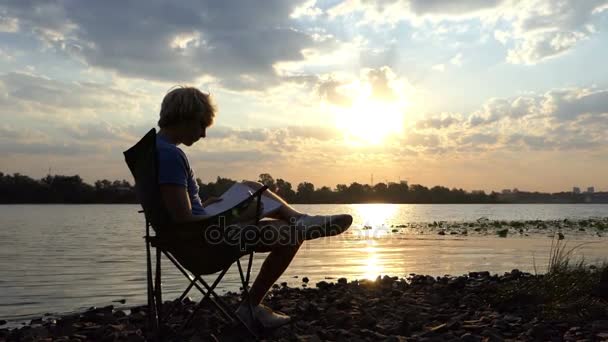 blonder Mann sitzt, liest und schreibt seine Notizen am Ufer des Flusses bei Sonnenuntergang - Filmmaterial, Video
