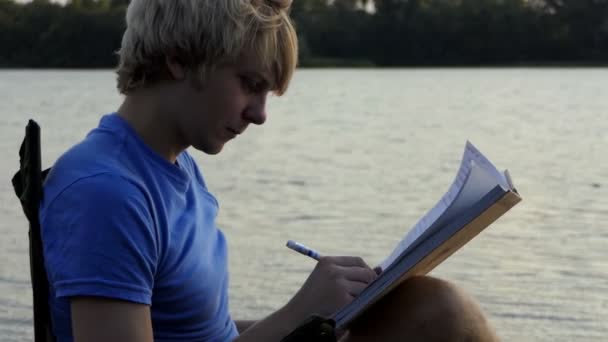 Muž s blond vlasy sedí, přečte a zapíše jeho poznámky na břehu při západu slunce v Slo-Mo - Záběry, video
