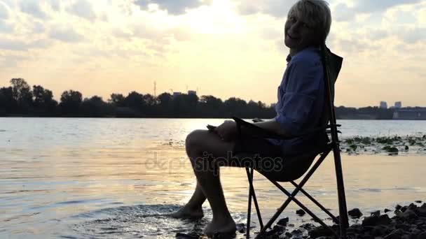 Nuori mies istuu ja hymyilee ja liikkuu paljain jaloin vedessä joen rannalla
 - Materiaali, video
