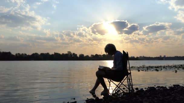 Yazar oturur, okur ve Slo-mo gün batımında notlarından bir nehir aşağı koyar - Video, Çekim