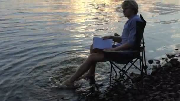 Ταλαντούχος άνθρωπος κάθεται και χαμόγελα και κινήσεις του ξυπόλητος σε νερό σε ένα Riverbank - Πλάνα, βίντεο