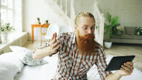 Young bearded man using tablet computer having video chat sitting in bed at home - 写真・画像