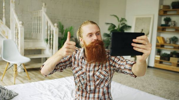 Young bearded man using tablet computer having video chat sitting in bed at home - Фото, изображение