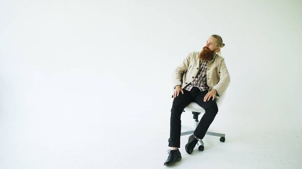 Серйозний бородатий чоловік сидить на поворотному офісному стільці на білому тлі
 - Фото, зображення