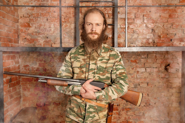 uomo magro con la barba in abiti militari e in piedi con una pistola contro un muro di mattoni
 - Foto, immagini