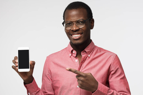 Happy african male isolated on grey background demonstrating blank smartphone and pointing to screen with expression of joy and interest. Copyspace for advertising products and services. - Foto, Bild
