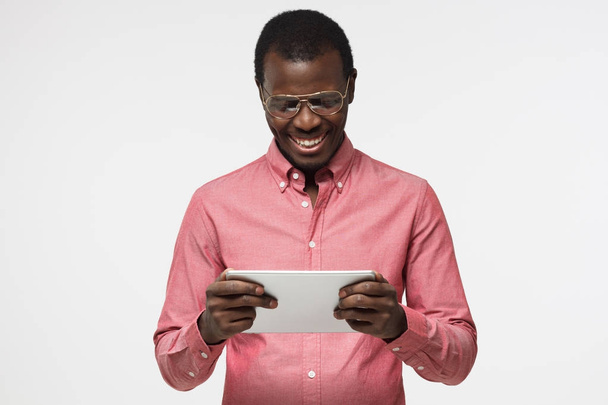 Indoor closeup of young handsome African man isolated on white background dressed in shirt, wearing big eyeglasses, looking at tablet screen with smile laughing while communicating or surfing net - Photo, image