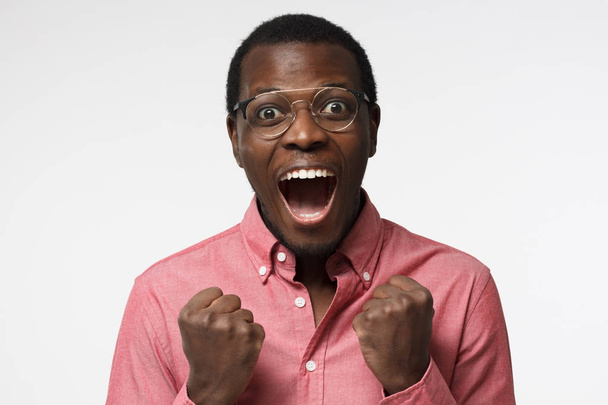 Young attractive black male in glasses shouting while his team win, raised both fist in victory gesture, isolated on gray background - Foto, Imagem