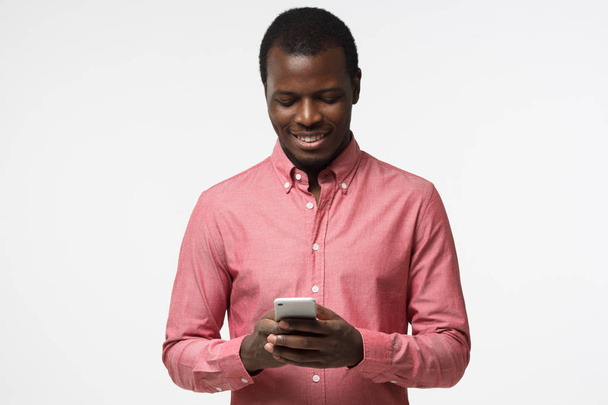 Closeup photo of casually-dressed African American male standing isolated on gray background looking attentively at screen of cellphone, browsing web pages and smiling nicely while chatting - Fotó, kép