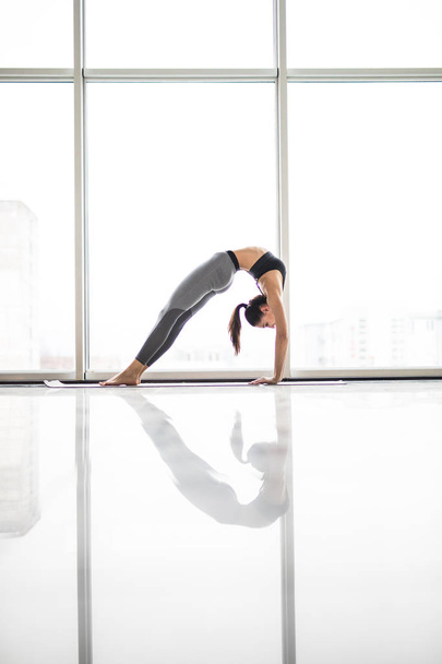 Jovem mulher atraente praticando ioga, alongamento em Elbow Bridge exercício, Urdhva Dhanurasana pose contra janelas panorâmicas
 - Foto, Imagem