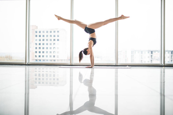 Young attractive yogi woman practicing yoga concept, standing in variation of Pincha Mayurasana exercise, handstand pose, working out, wearing sportswear, full length against windows - Foto, imagen