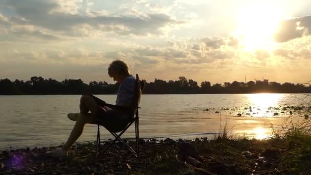 Okos ember ül a folyóparton, írja a naplemente összecsukható szék - Felvétel, videó