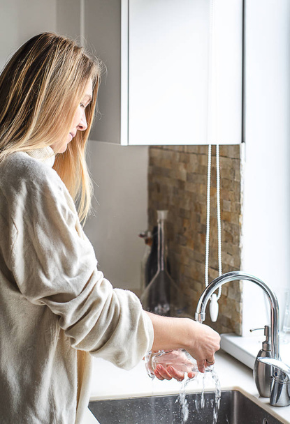 Woman with long blonde hair washing dish or pouring glass with running fresh drink water at kitchen faucet - Photo, image