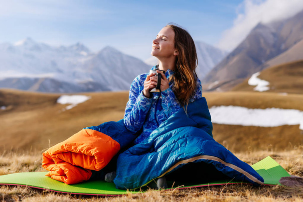 happy young girl sits in a sleeping bag on the background of the Caucasian mountains, enjoys nature and clean air, meditates - Fotó, kép