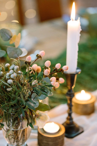decoration, planting, wedding concept. on the table there is small posy with little berries of light pink colour and various leaves, it is standing by candles in different holders, small and big - Photo, image