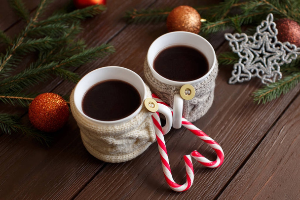 Зимняя любовь. Чашки кофе или чая с вязаными держателями чашки возле рождественских конфет и рождественский торт на деревянном фоне с копировальным местом. Начало рождественского утра
 - Фото, изображение