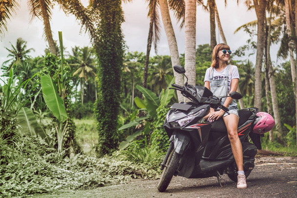 Young happy woman on a motorbike in the jungle rainforest of a tropical Bali island, Indonesia. Freedom concept. Lady on a scooter. - Фото, изображение